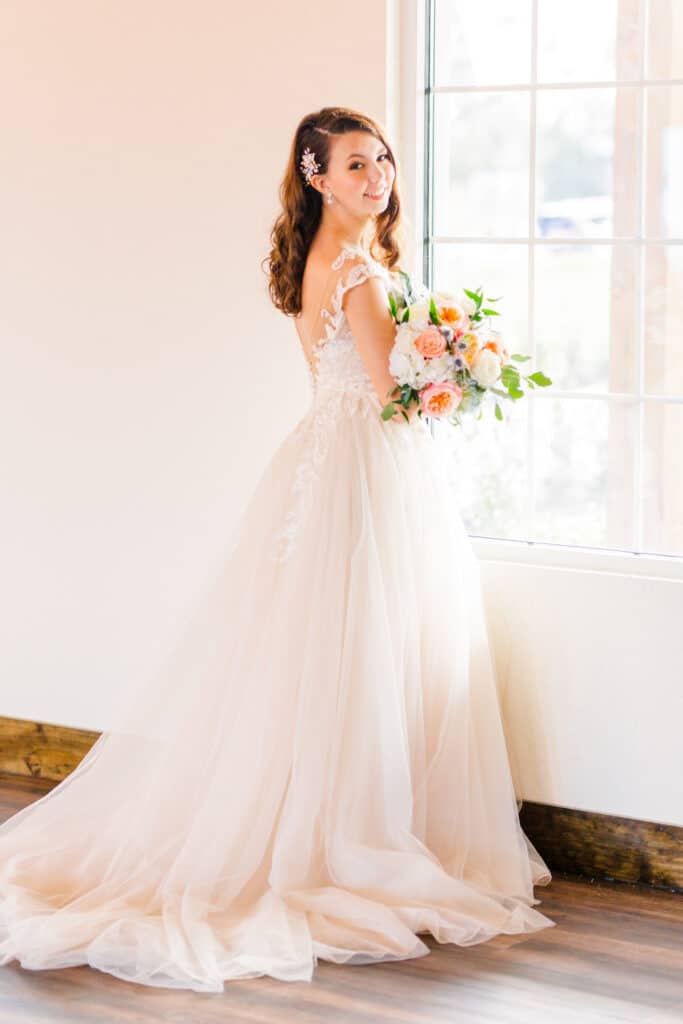 bride stands facing window in wedding dress holding bouquet smiling back at the camera