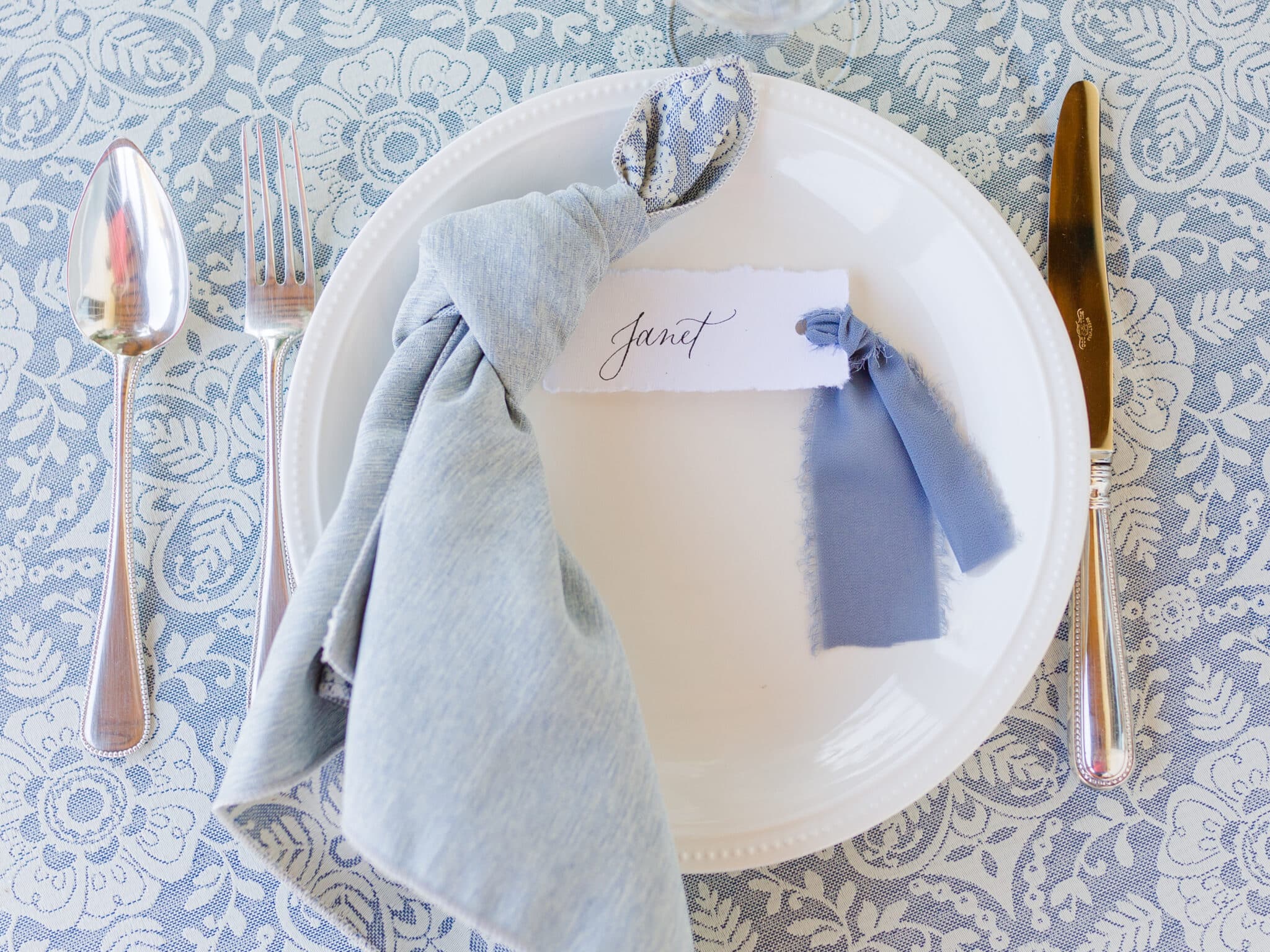 white plate with light blue napkin tied in a knot sittign on top of it with hand calligraphy place setting card