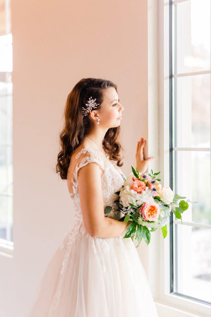 woman in wedding dress holding flower bouquet looks out window and gently places hand on window frame