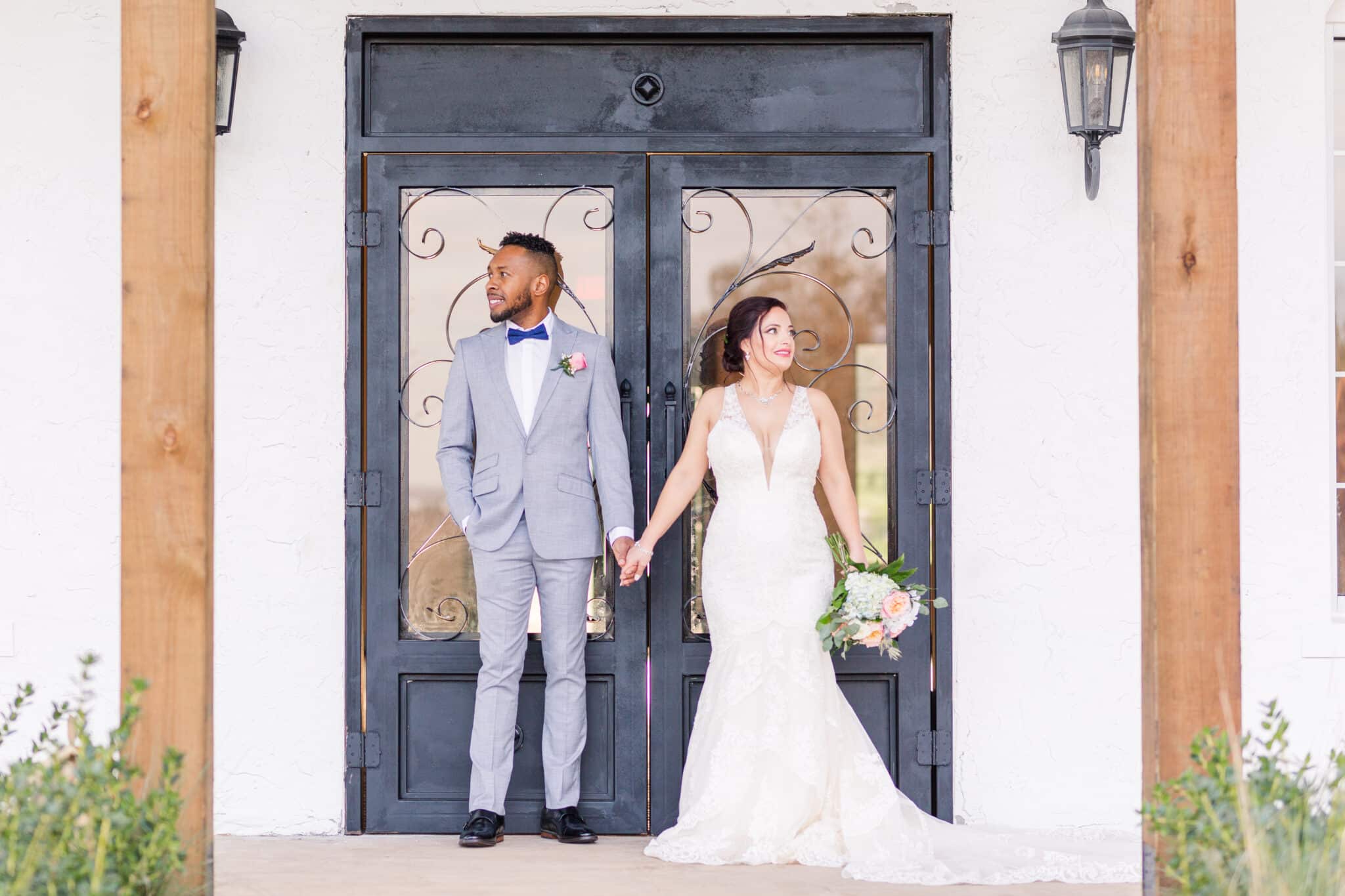 couple stand in front of black doors of wedding venue holding hands and looking off to the side away from each other in suit and wedding dress