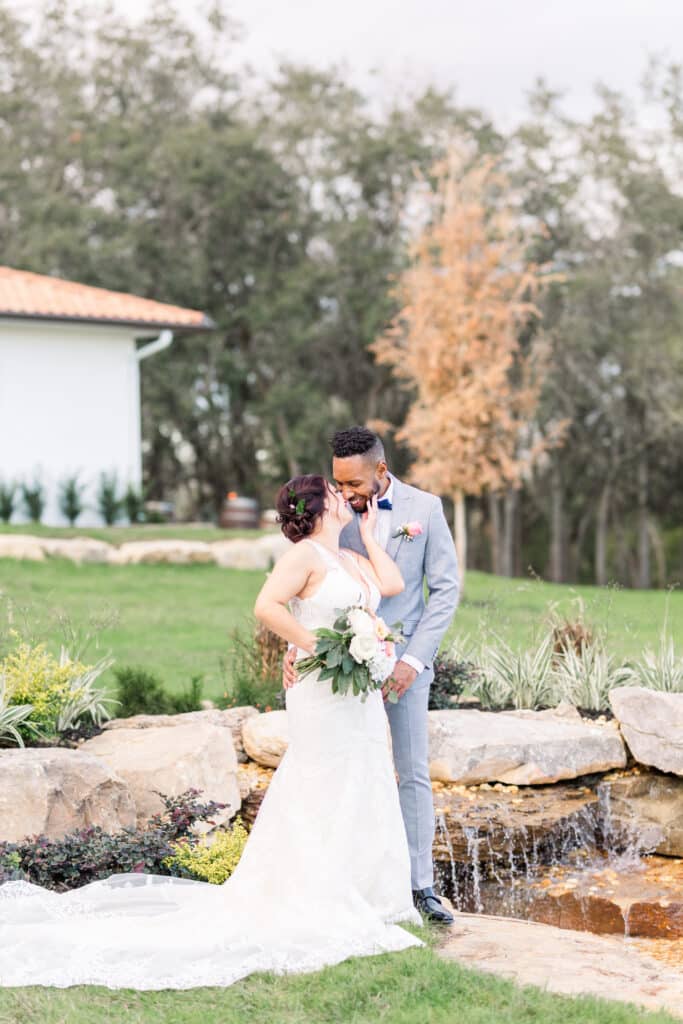 couple stand together in front of waterfall and rock landscaping outdoor kissing on wedding day