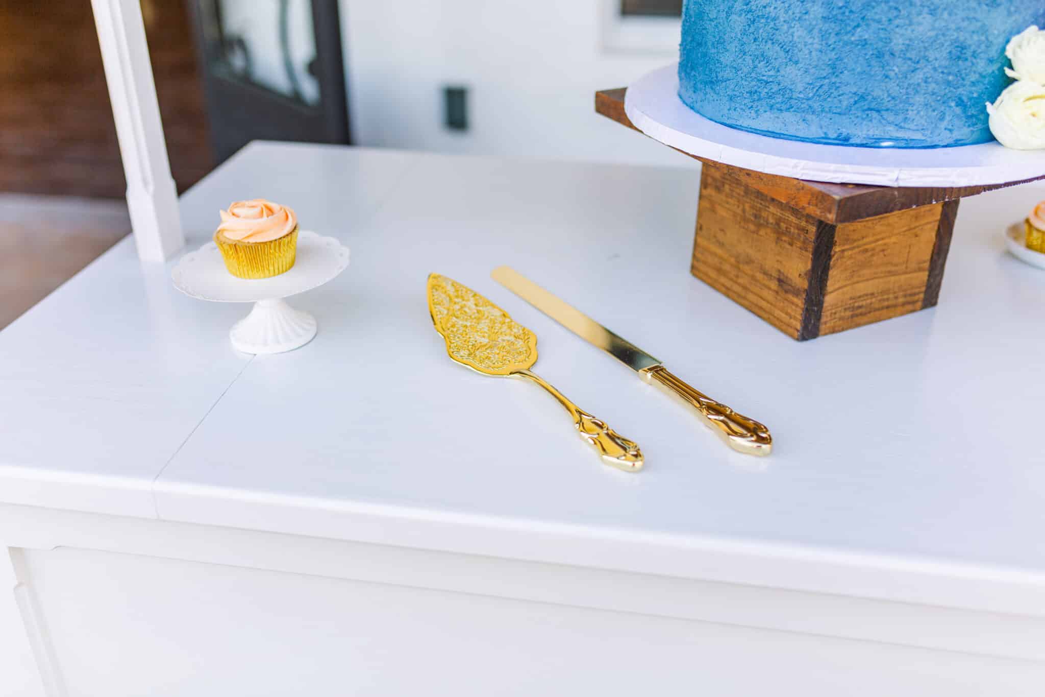 close up of cake cutting set sitting on the table next to a single cupcake on a tiny stand and a wooden cake stand next to it