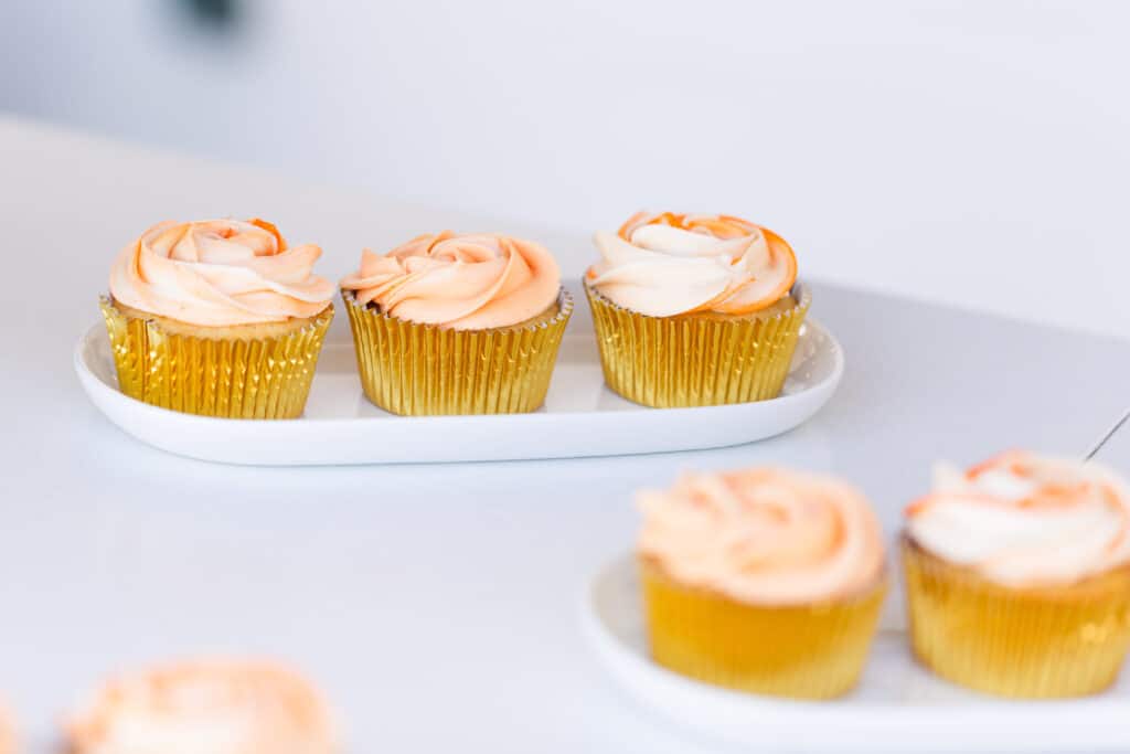 five cupcakes with golden cupcake liner and pink frosting in a rose design sit on white surface