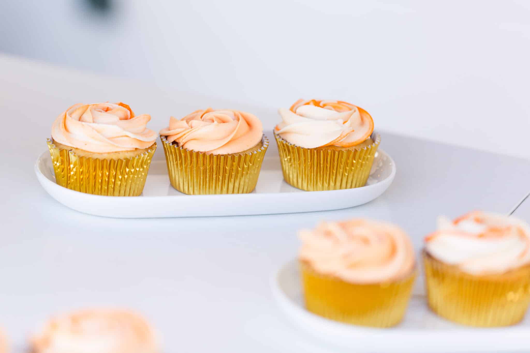 five cupcakes with golden cupcake liner and pink frosting in a rose design sit on white surface