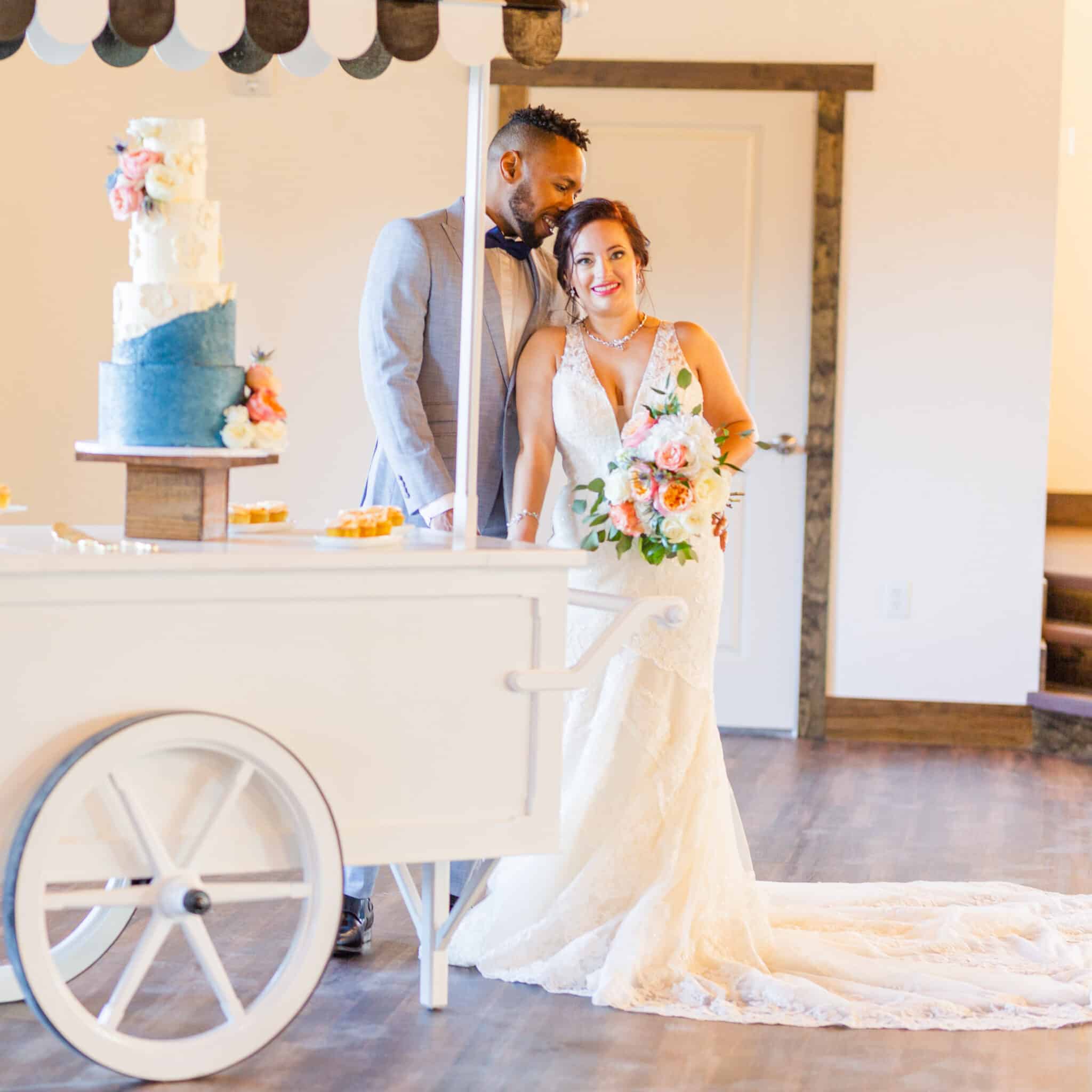 man stands next to woman in wedding dress holding bouquet as he kisses her head and a cart with wedding cake is to their left