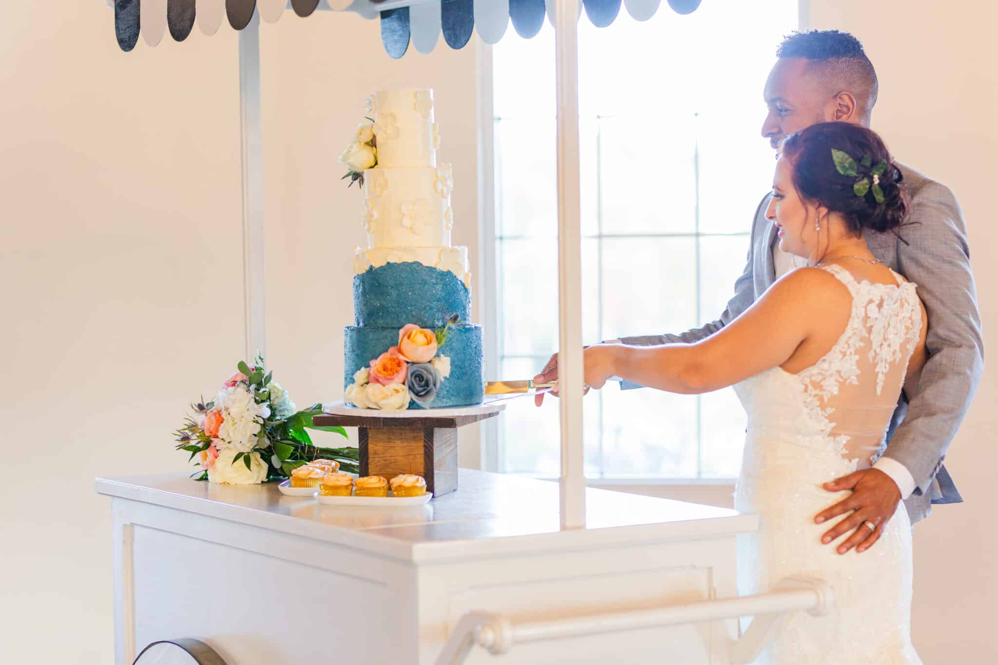 couple stands together cutting wedding cake decorated with flowers and blue and white icing