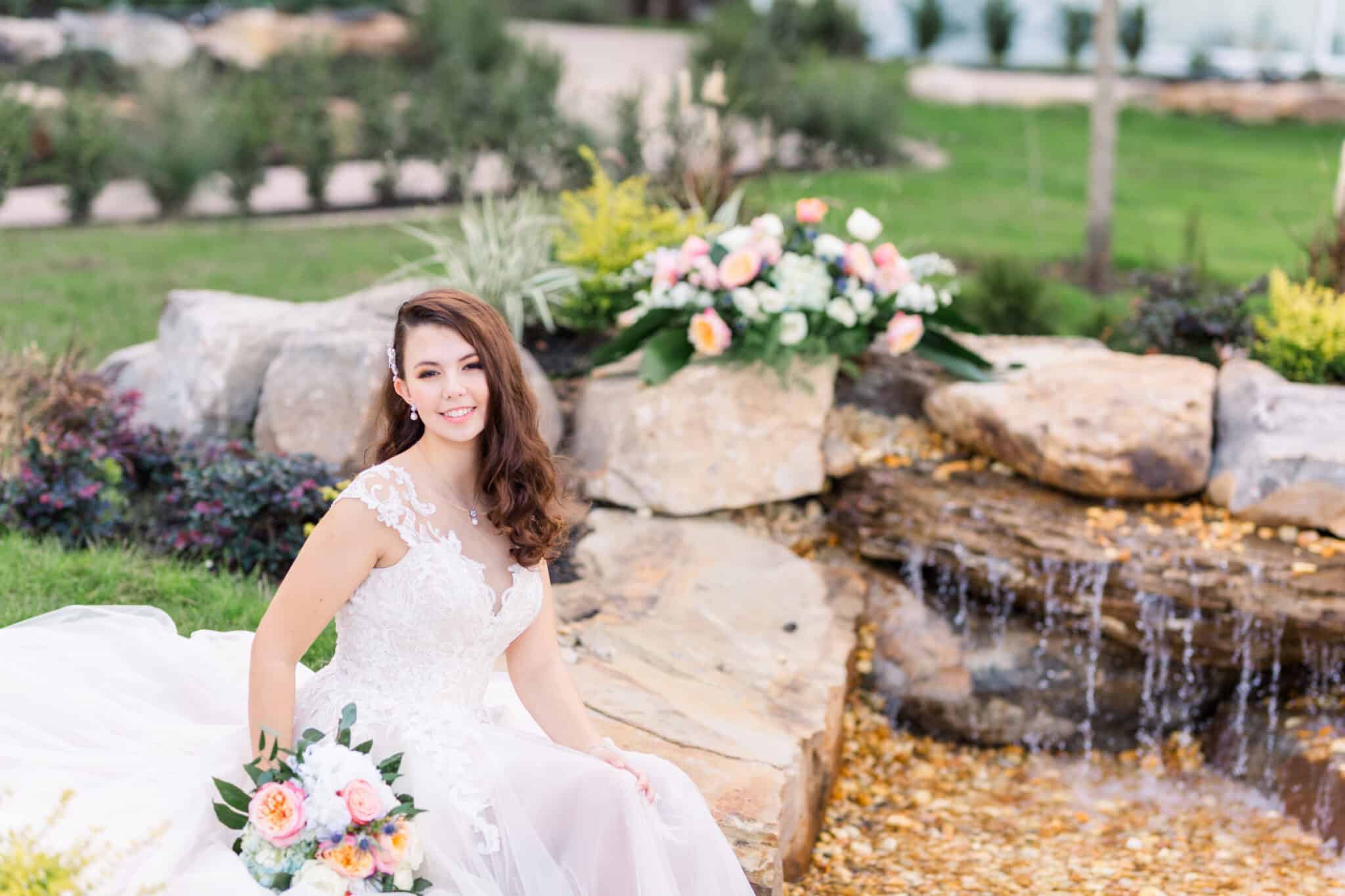 bride sits on rocks near waterfall landscape in wedding dress with flower bouquet