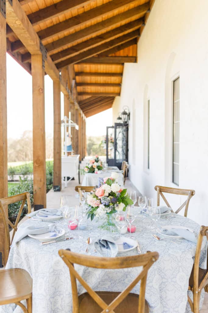 table with cross back wooden chairs set for event outside under awning of venue with floral centerpiece