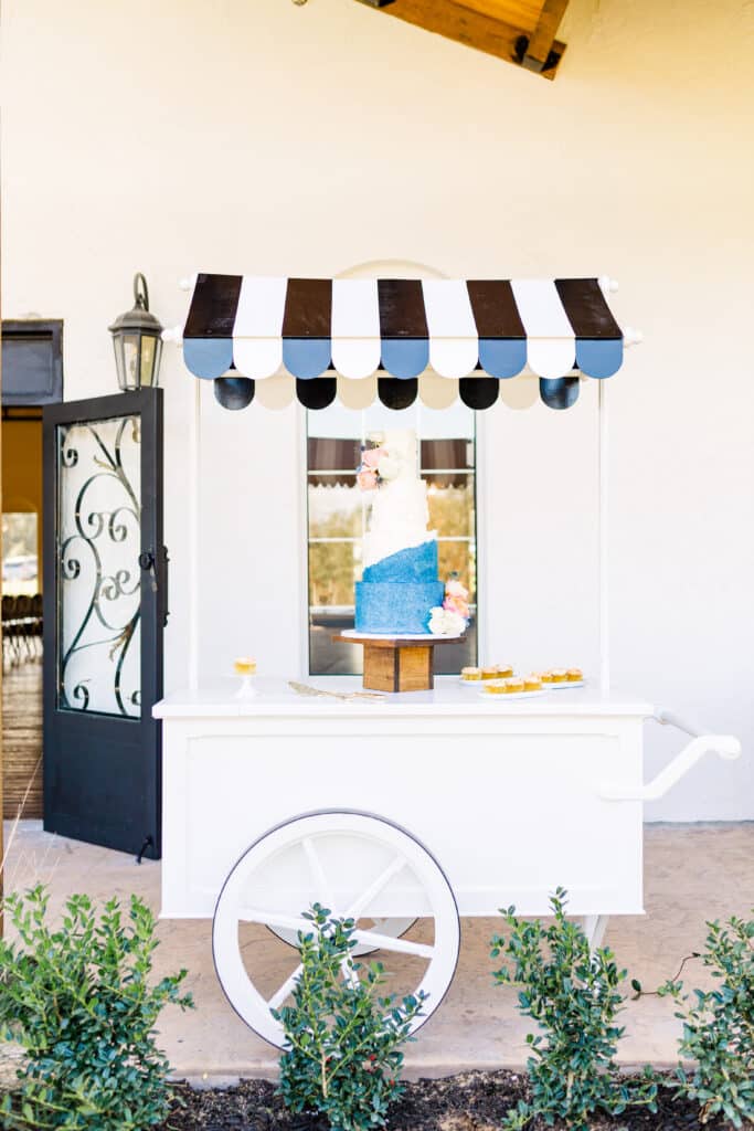 white cart with black and white awning over it has cake sitting on it with blue and white design in front of a window