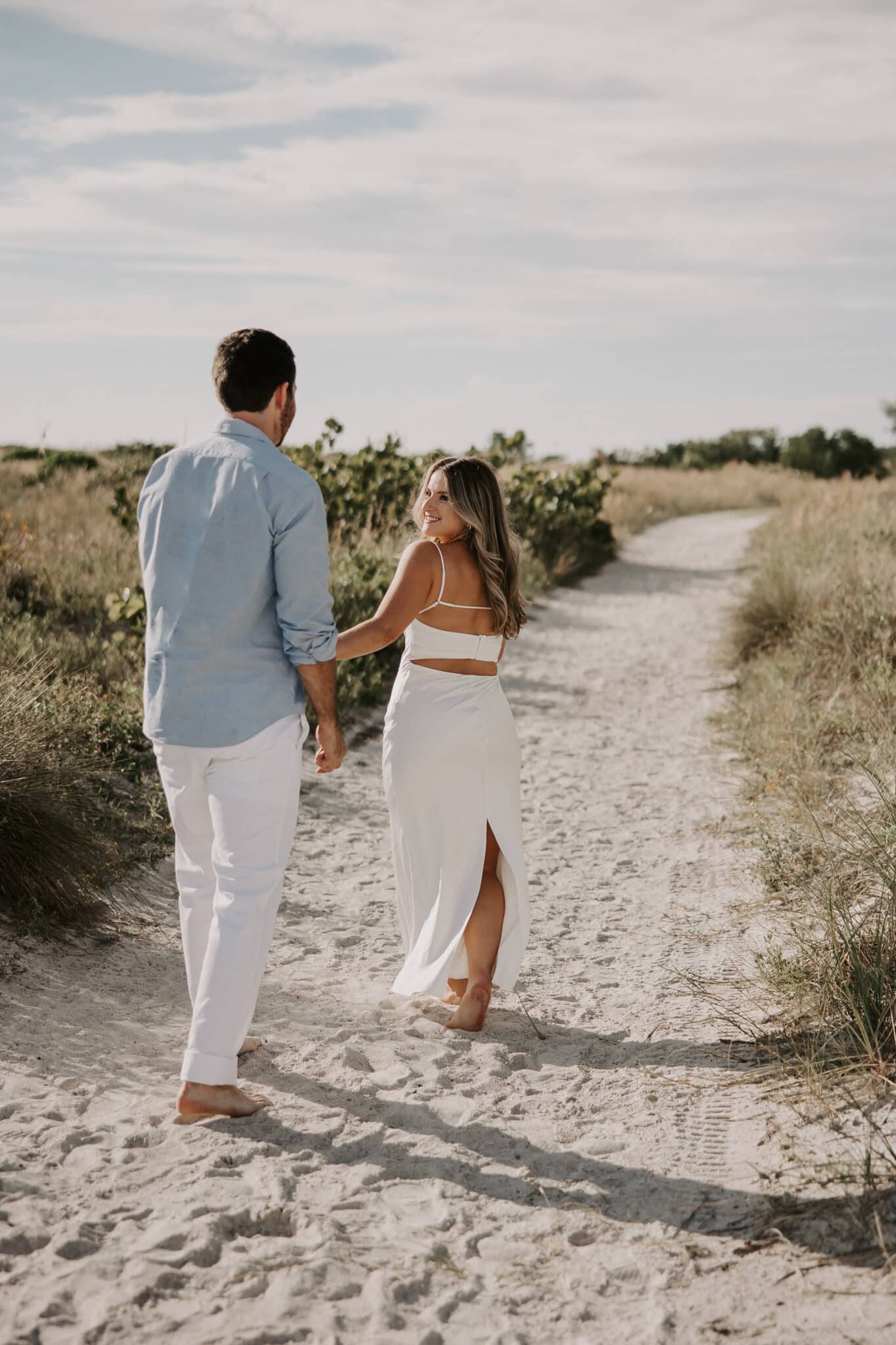 woman walks along sandy path in white dress while looking back at man walking behind her