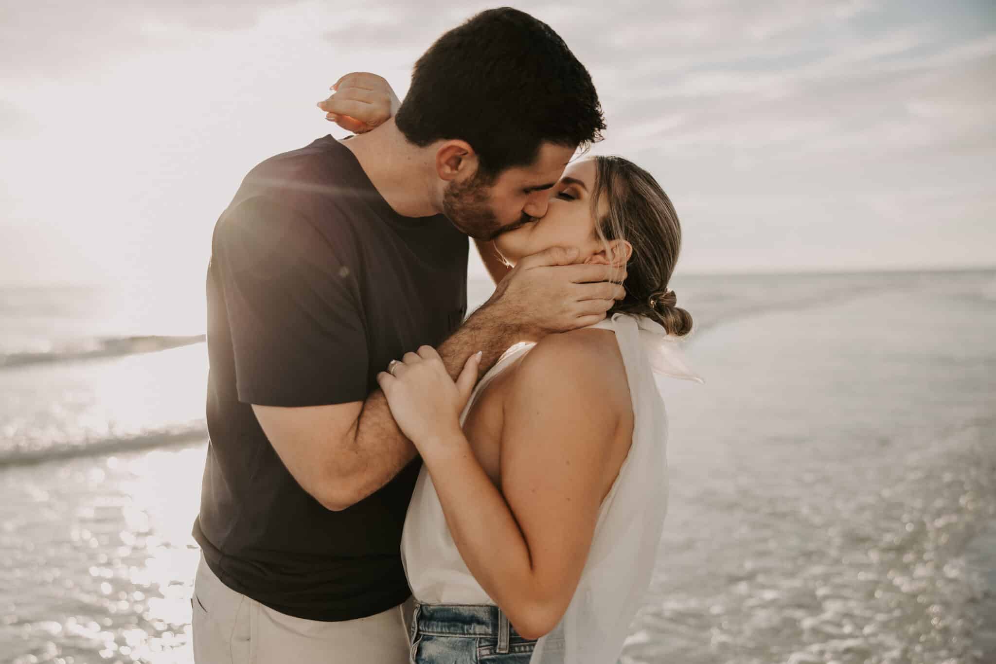 engaged couples kiss with waves of the ocean crash behind them