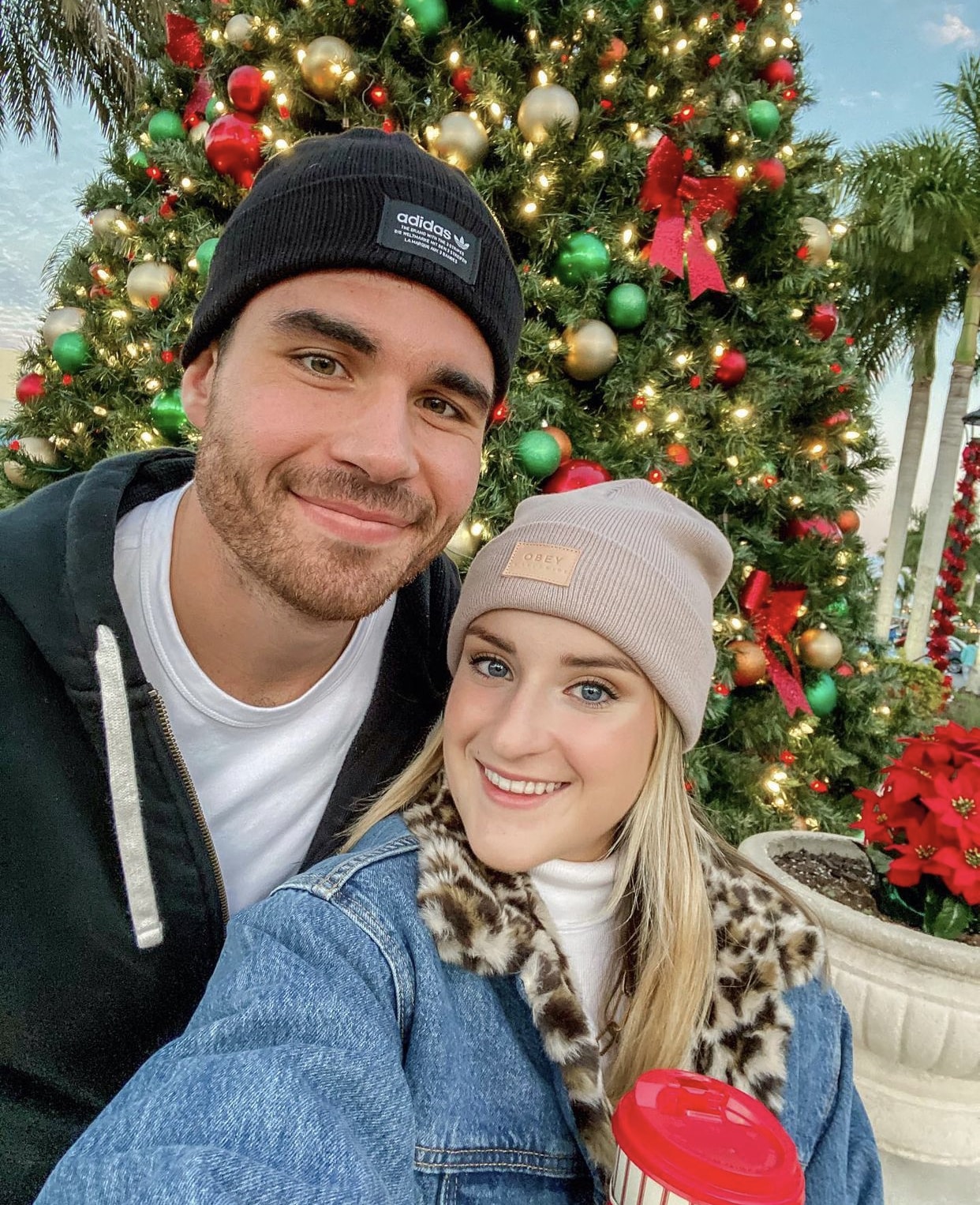 man and woman stand together taking a selfier wearing hats and jackets in front of decorated christmas tree