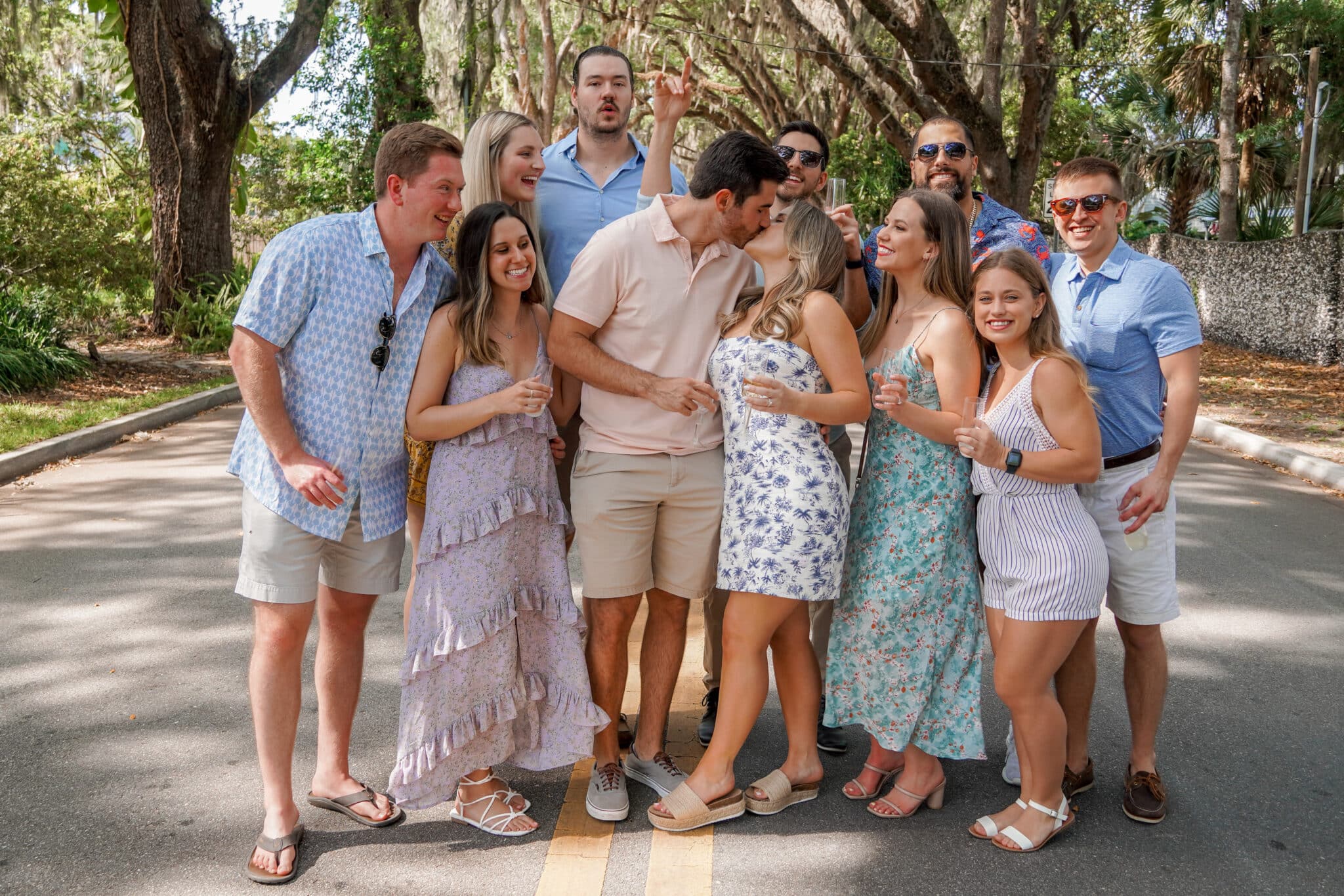 couple stand in the middle of a bunch of family and friends kissing as they all hold champagne glasses in the middle of the street