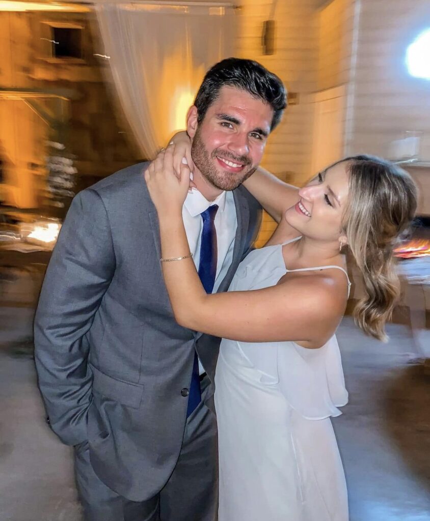 girlfriend holds around the neck of boyfriend wearing suit and tie smiling at camera