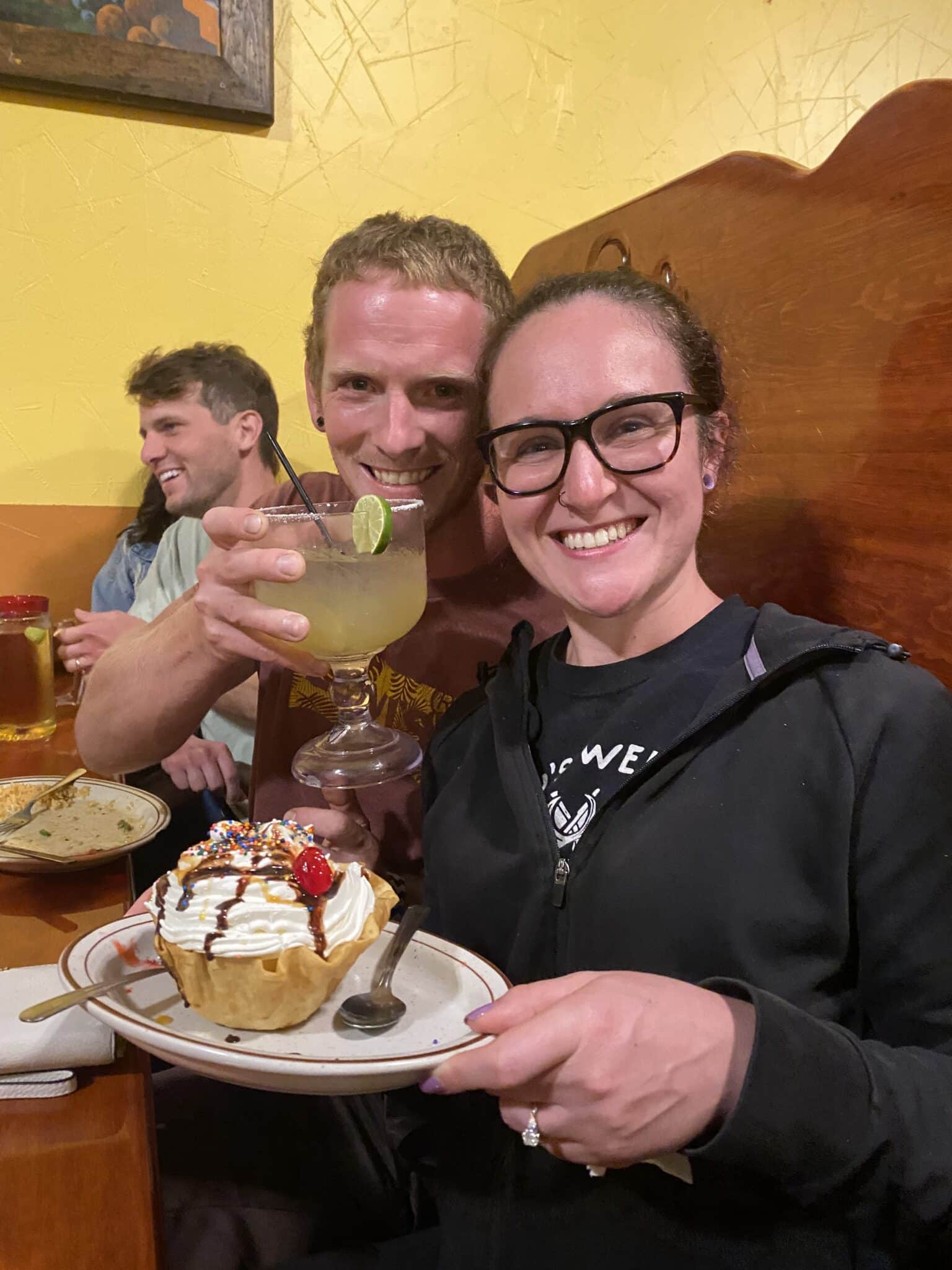 woman with glasses wears hoodie and holds dessert on plate while man sitting next to her smiles holding a drink