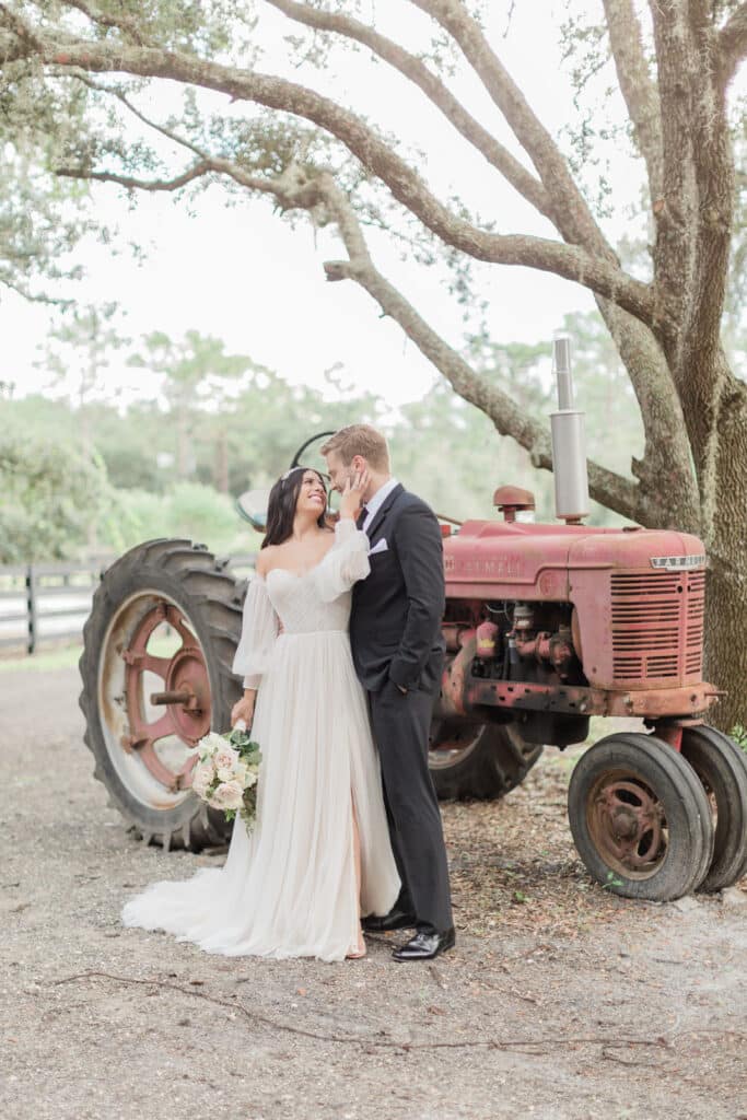 bride and groom standing in front of vintage red tractor at The Barn at Hidden Oaks Farm of Lake Mary
