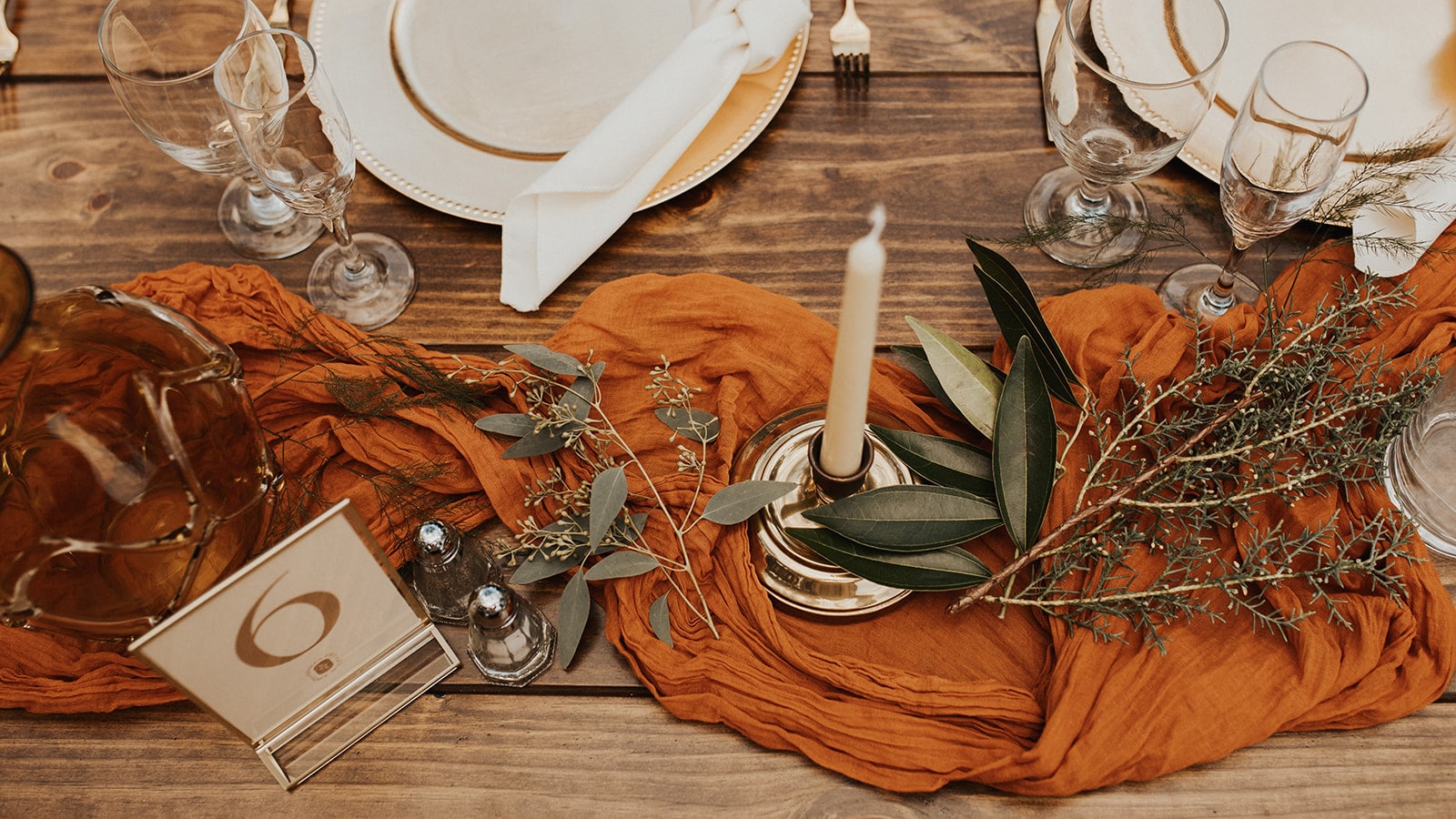 overhead view of tablescape with burnt orange cheesecloth down the center of the table and small sprigs of greenery with candlesticks and a table number to the left