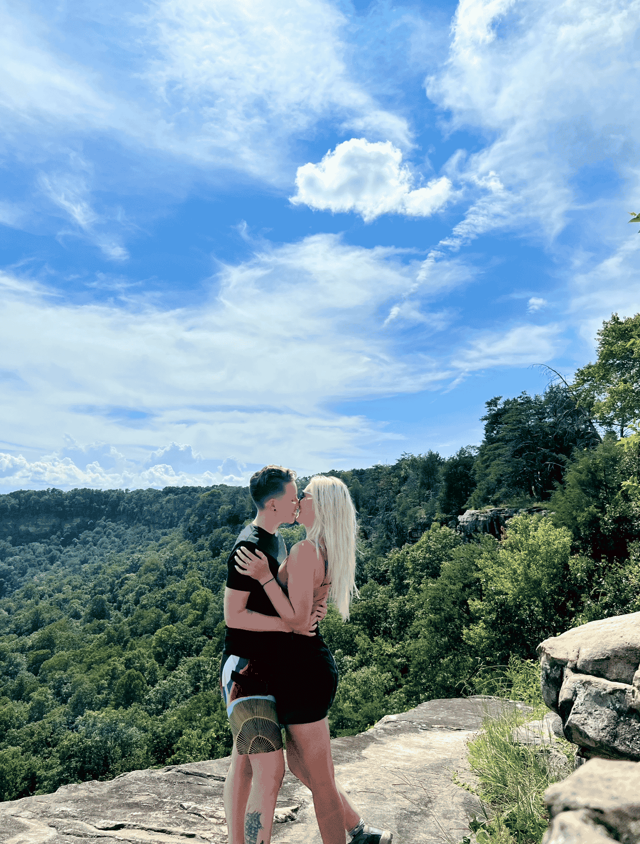 couple stands kissing on mountain top ledge overlooking treetops