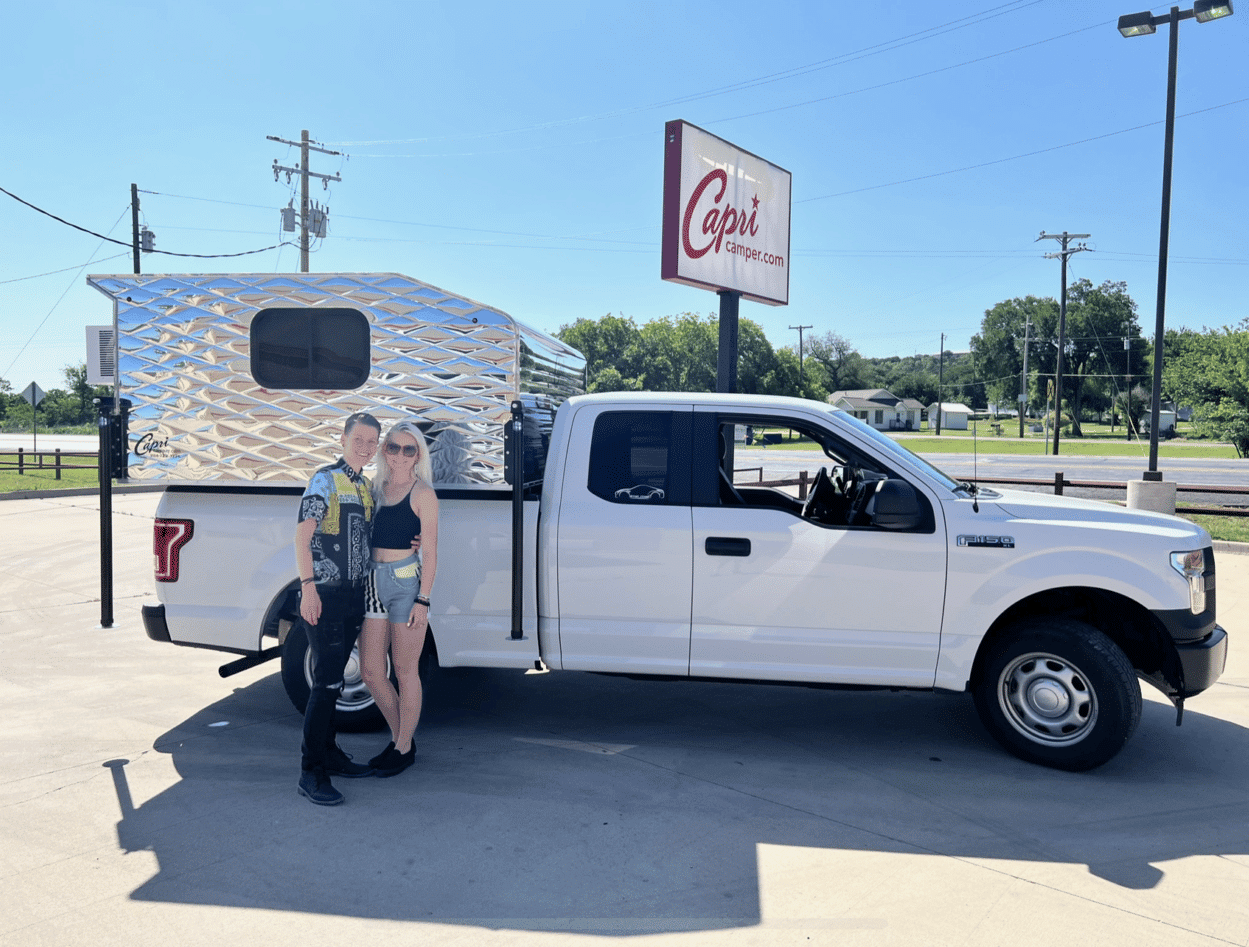 two woman stand together in front of white pickup truck with a camper attached to the bed of the truck