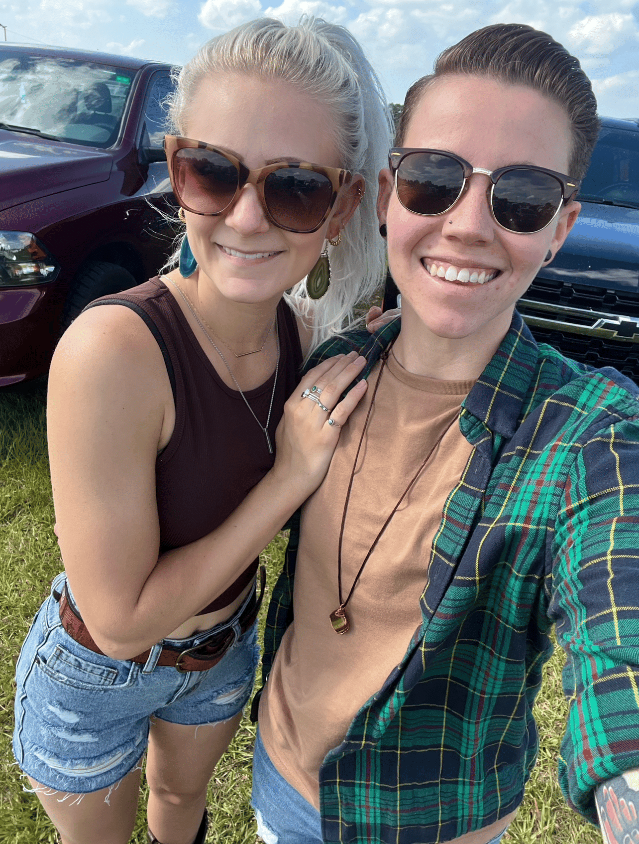 couple wearing sunglasses stand together taking a selfie in front of parked cars behind them