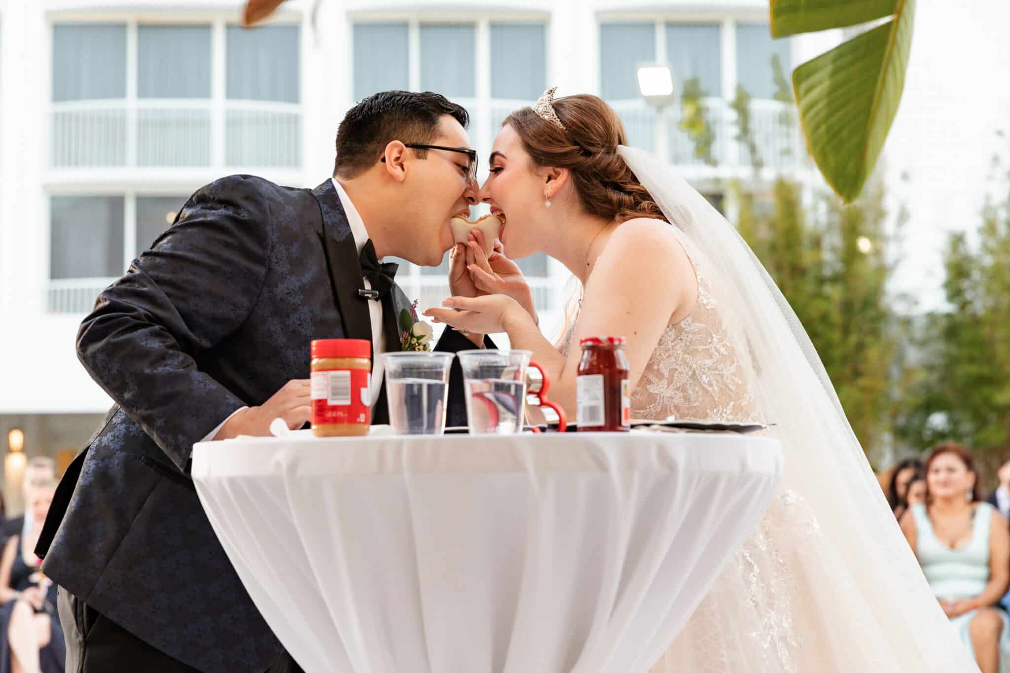 bride and groom leaning in to take a bite of a peanut butter and jelly sandwich next to cocktail table during wedding day