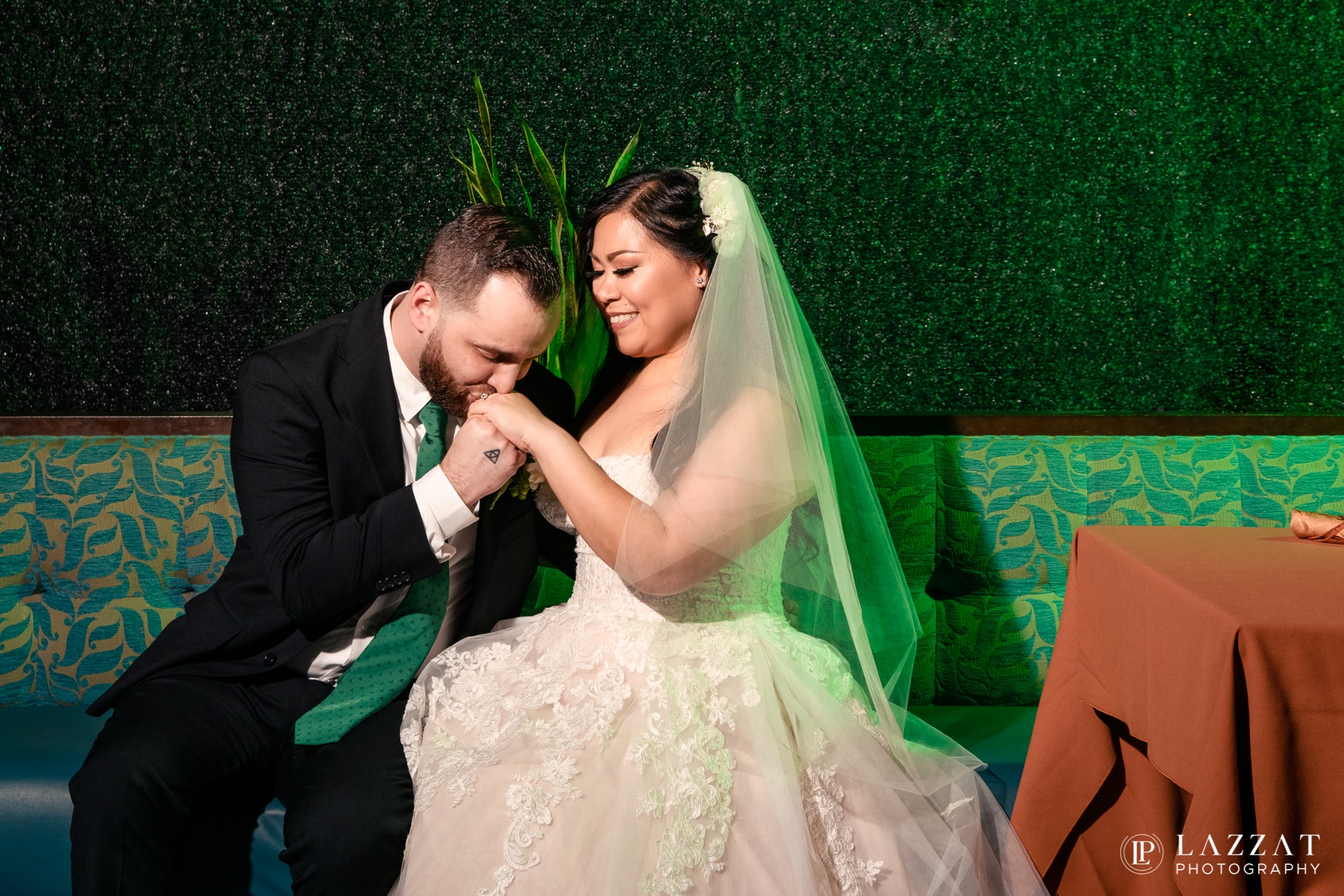 groom leans over and kisses brides hand as they're seating at booth with green backlight