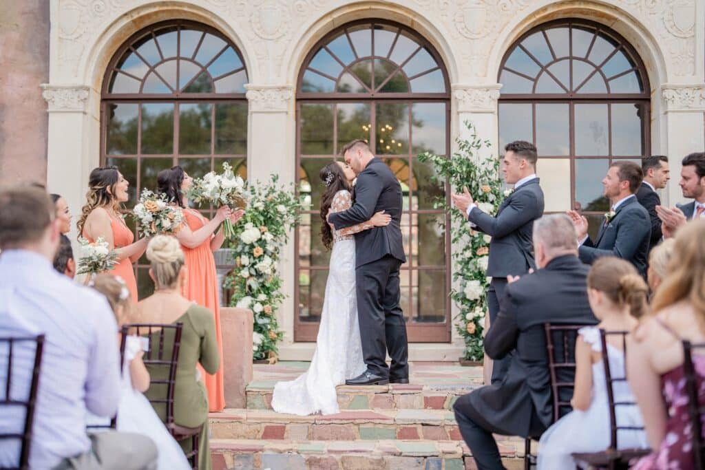 bride and groom kissing on the steps of an outdoor wedding with three arched windows as a backdrop