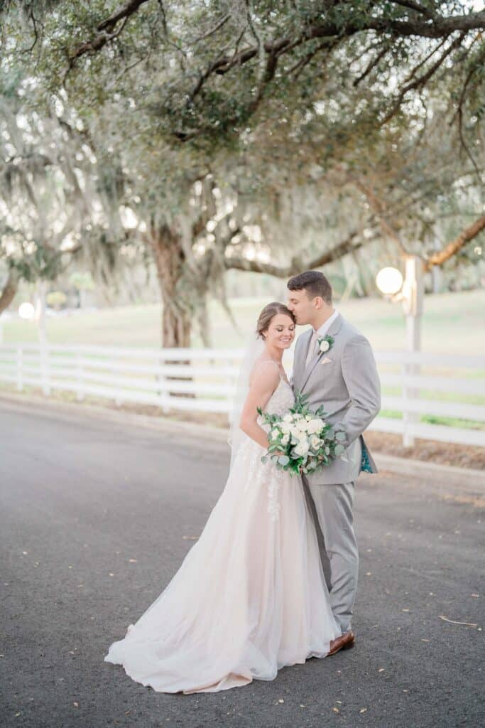bride and groom in grey suit on an asphalt road with a white wooden fence and trees in the background
