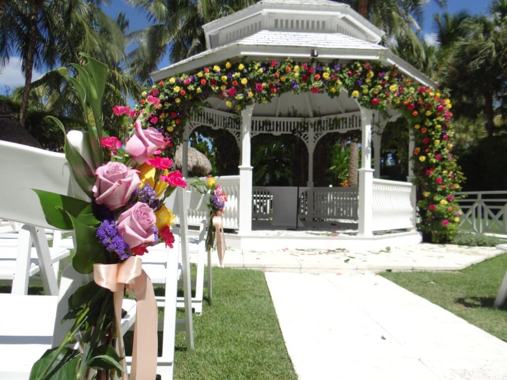 outdoor wedding ceremony aisle with lavendr and hot pink flowers at the end and a white runner leading up to the swag draped pergola