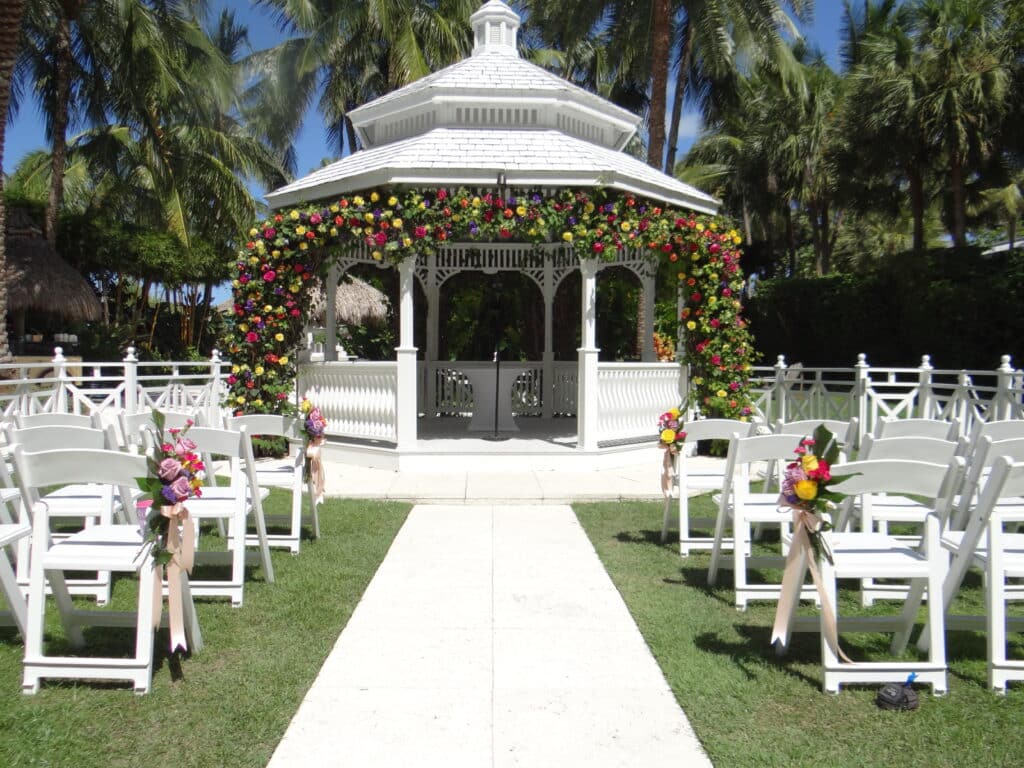 outdoor wedding ceremony with white runner leading up to southern outdoor pergola draped with green swag with colorful flowers