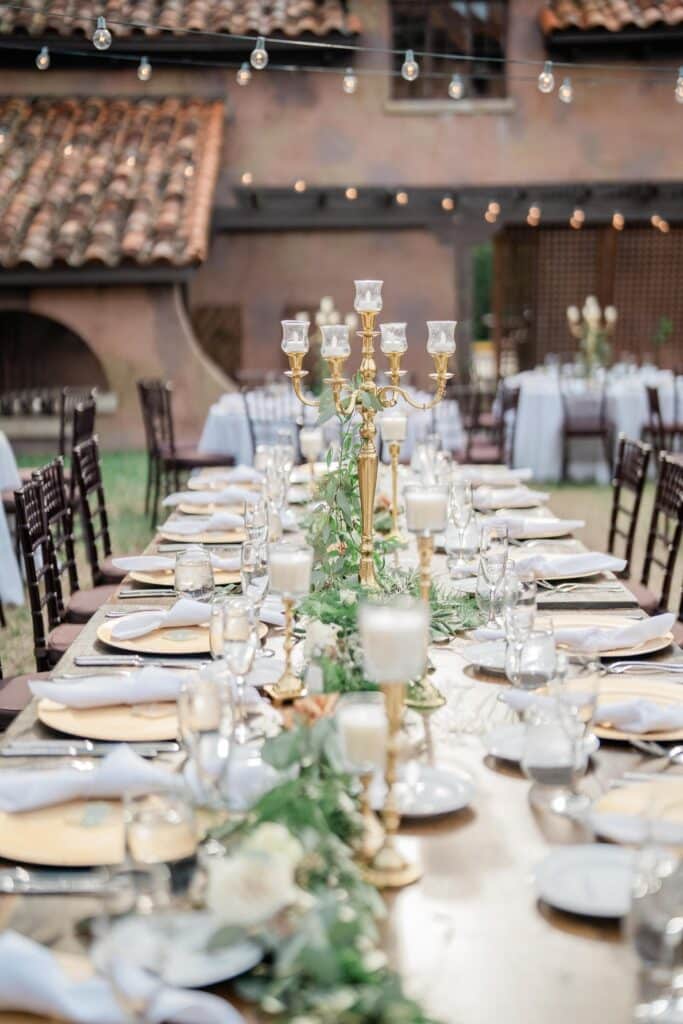 long table set with gold chargers for wedding reception at an outdoor venue with market lights and candleabras