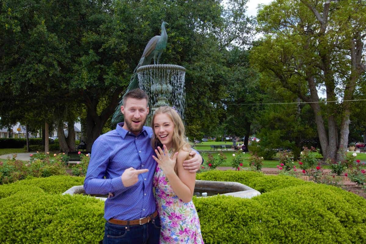 man stands holding girl pointing to her outstretched hand with new engagement ring in front of fountain outside