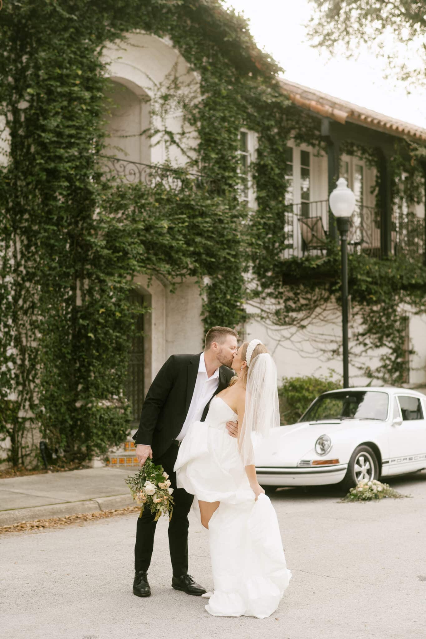 couple kiss for engagement picture outside of building with ivy on the walls and vintage car in the background