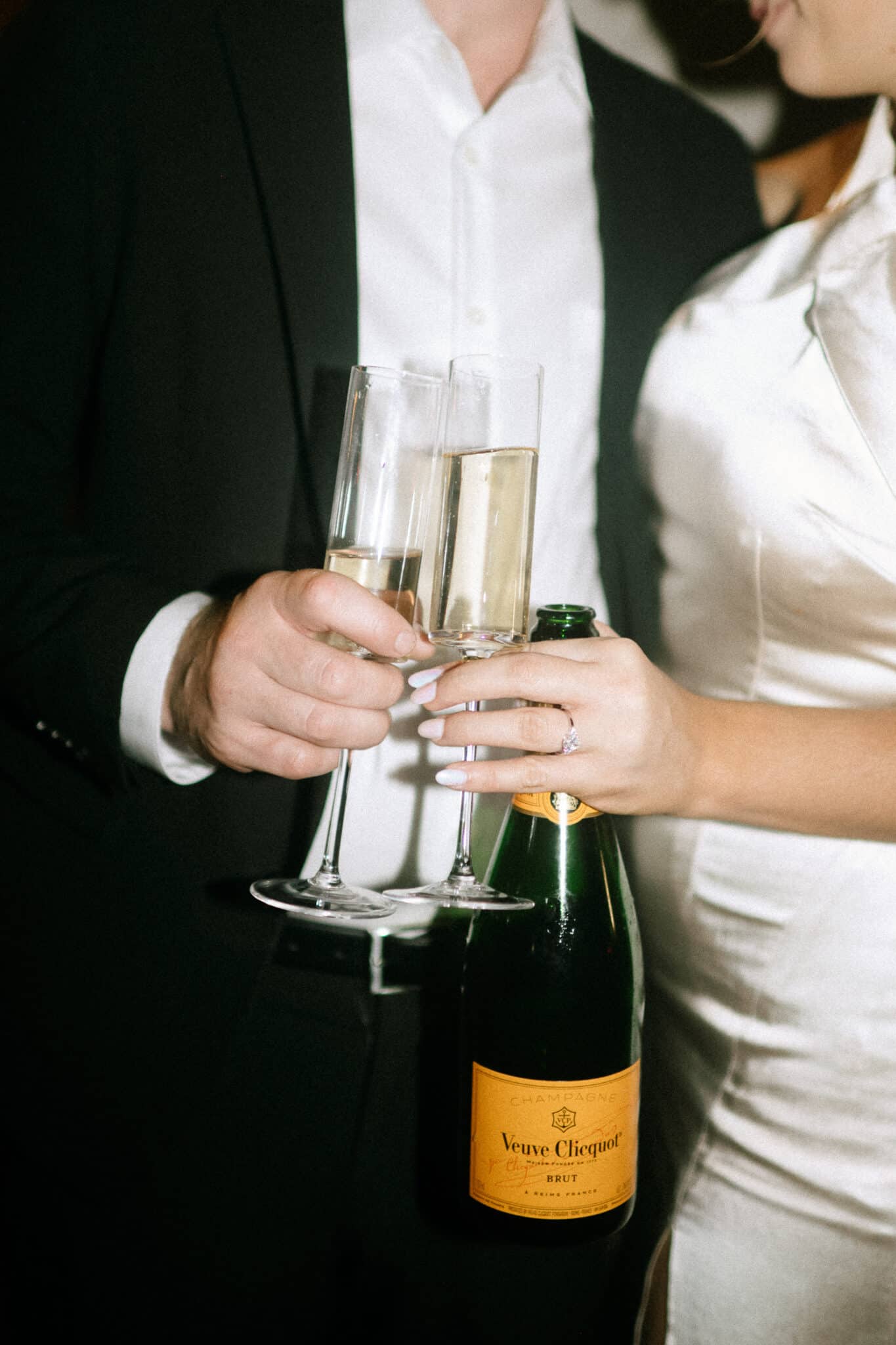 close up image of engaged couple holding glasses of champagne together with bottle of champagne behind the glasses