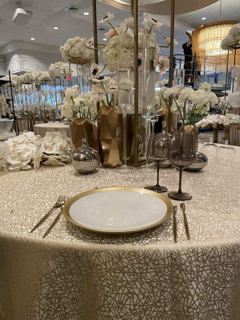 table sert with white trimmed with gold plates, gold flatware, gold linen and large centerpieces from Design One USA