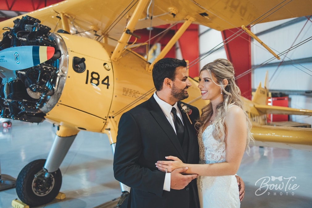 bride and groom smiling at each other in front of yellow bi-plane in a hangar at The Warbird Air Museum