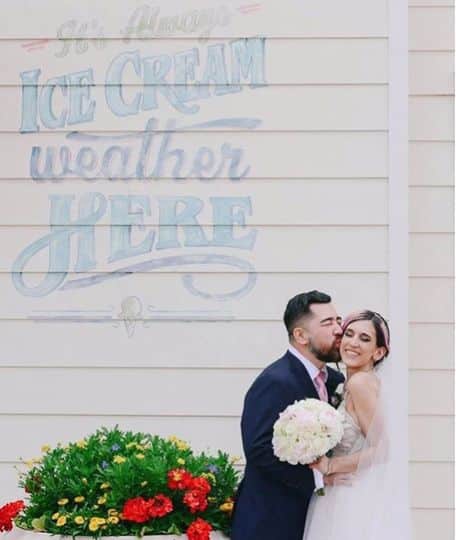 groom kissing bride as they pose for photo against old wall mural for ice cream shop at wedding coordinated by Fairy Tales & Wedding Bells