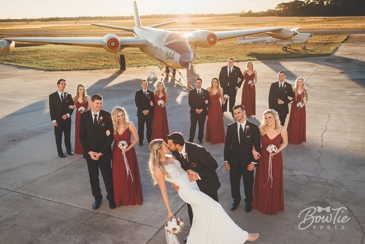 groom dips bride for a kiss in front of wedding party standing in front of a glider outside The Warbird Air Museum