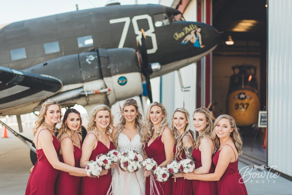bride and bridesmaids dressed in red in front of vintage airplane outside a hangar at The Warbird Air Museum