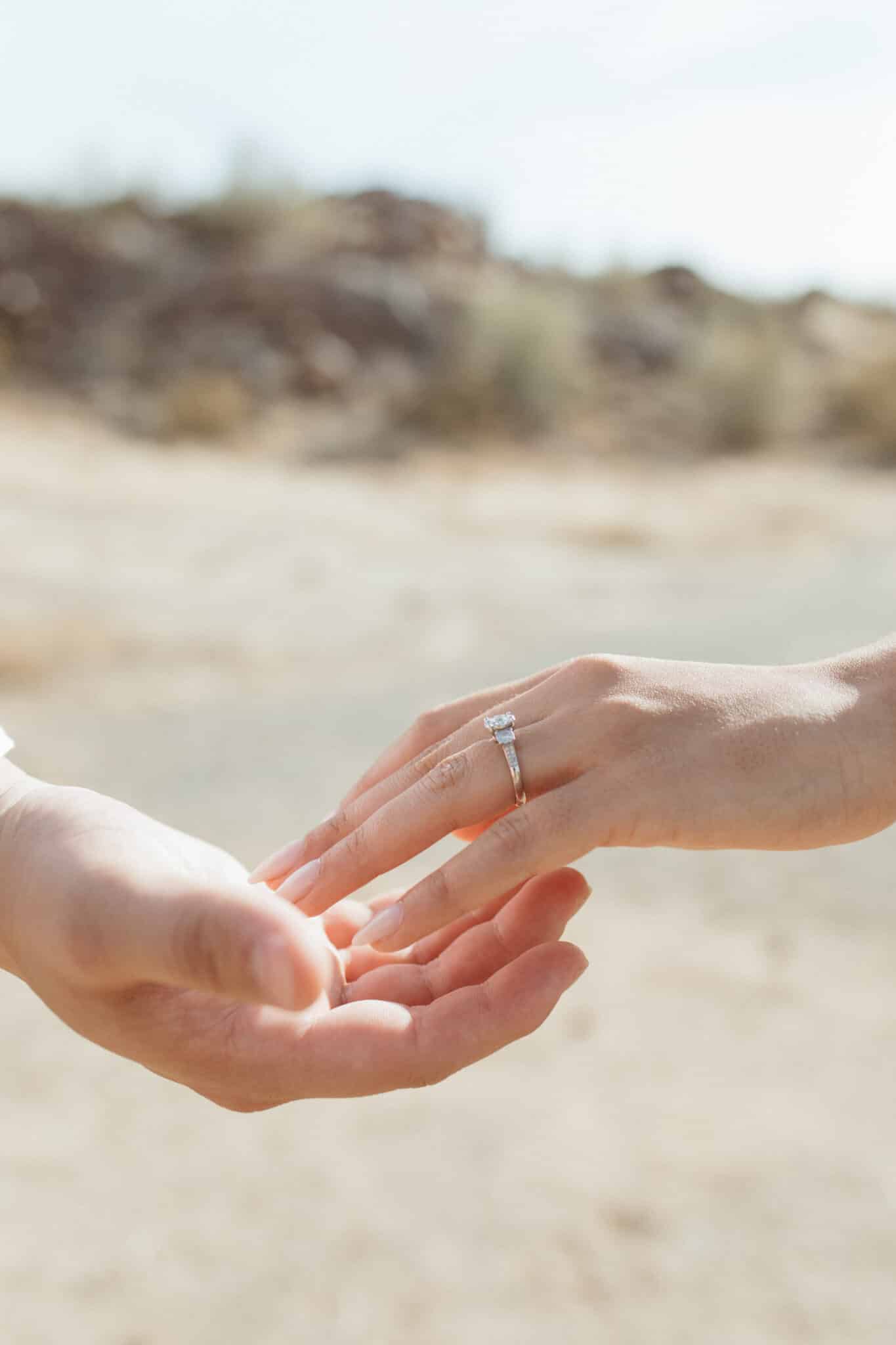 two hands lightly touching each other at the beach with sand dunes in the background and engagement ring on one of the hands