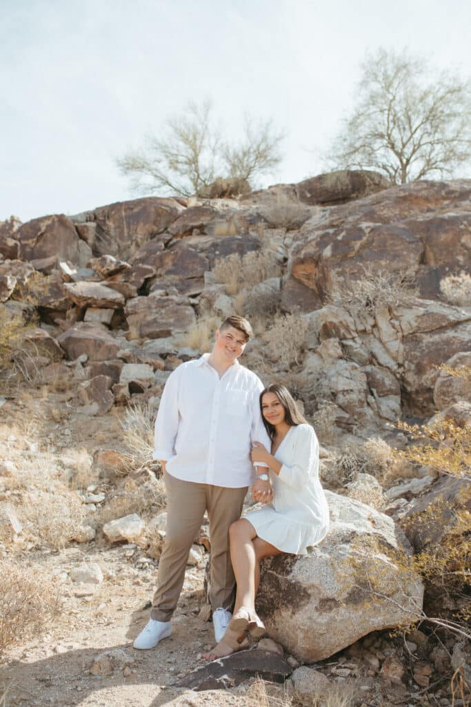 girl sits on boulder clinging onto mans arm as he stands next to her both wearing white