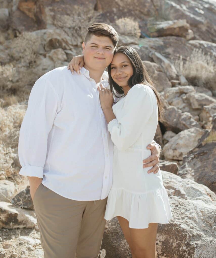 man wraps arm around the waist of woman wearing cream colored dress both smiling for a picture in front of many rocks