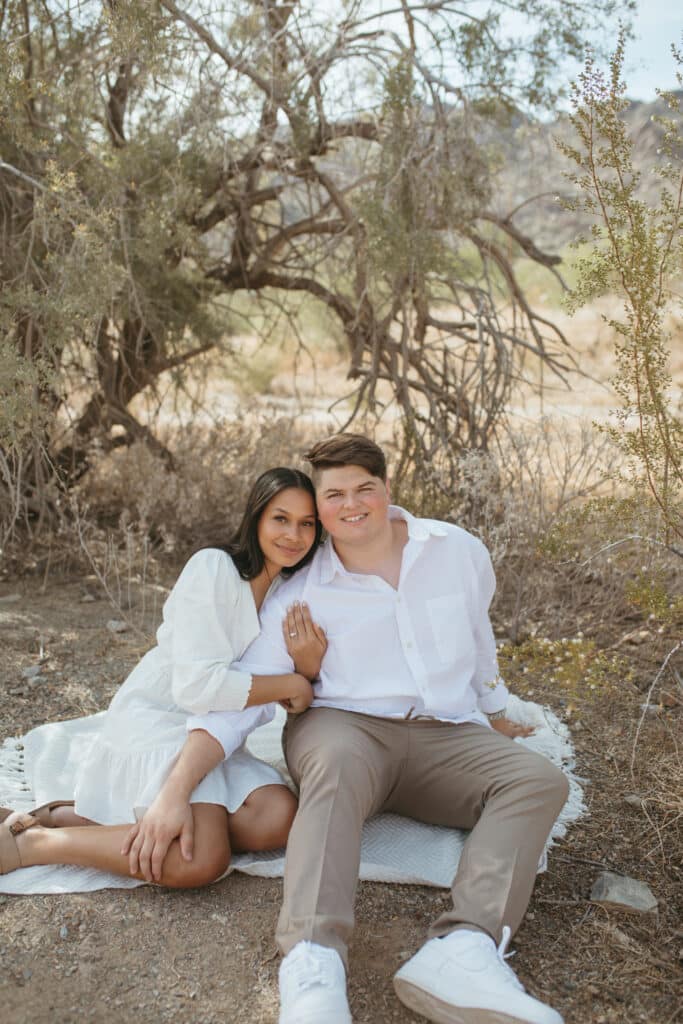 couple sits together posing for picture on blanket outdoor with unique shaped tree behind them