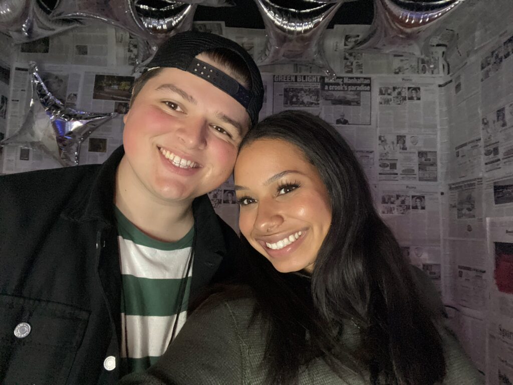 woman smiling poses next to man smiling with hat on backwards in front of newspaper covered wall behind them