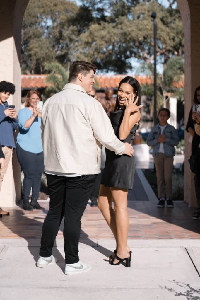 man stands next to girl facing the camera holding up her new engagement ring on her hand and smiling with her mouth open
