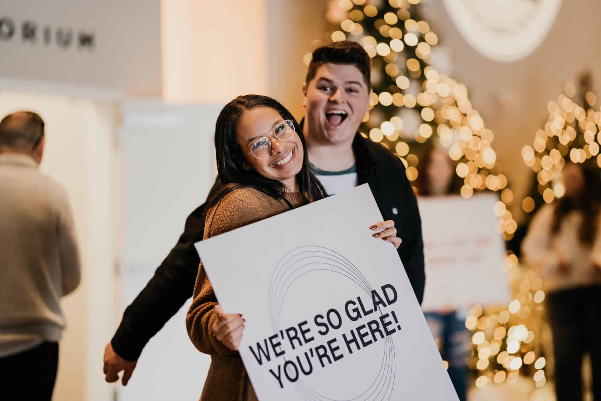 woman holding a sign that says we're so glad you're here smiles while man has goofy smile behind her