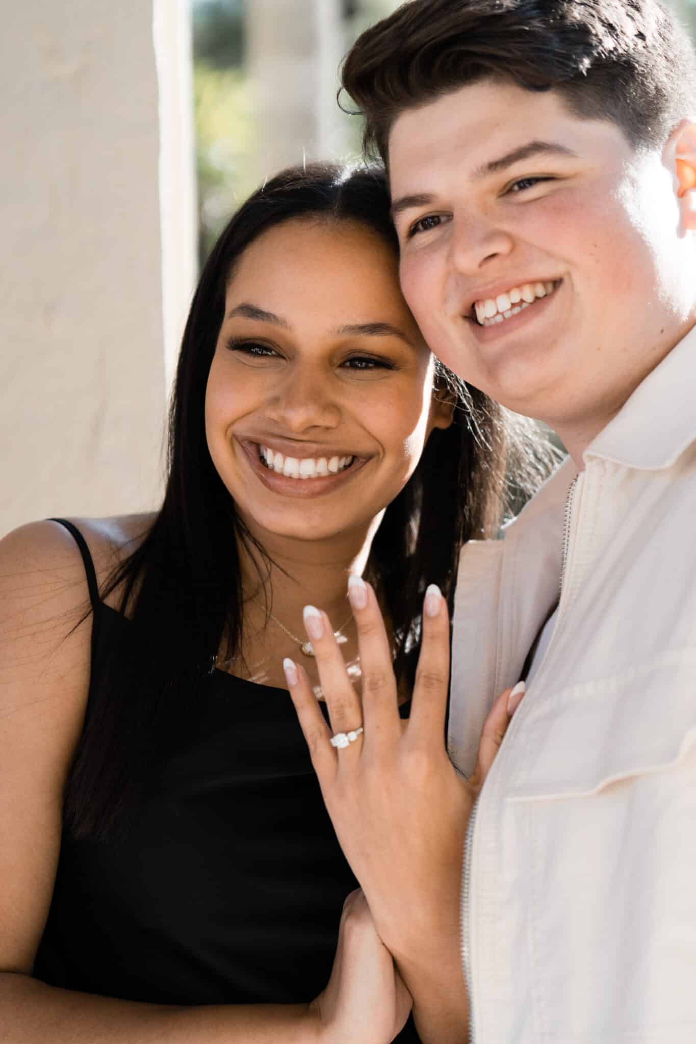 couple stand together smiling big while girl holds up hand in front of them showing her new engagement ring off