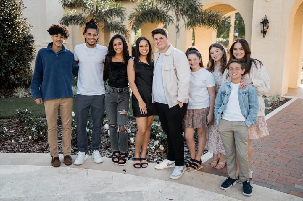 a group of kids and young adults stand together posing for a picture outside in front of palm trees and arched structure on a building