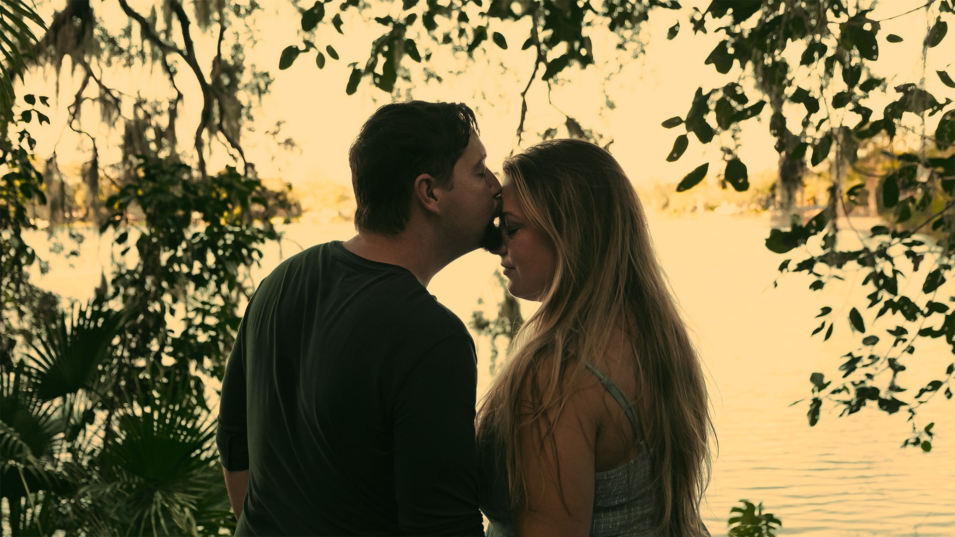 man leans over and kisses woman on forehead with body of water behind them and greenery surrounding them