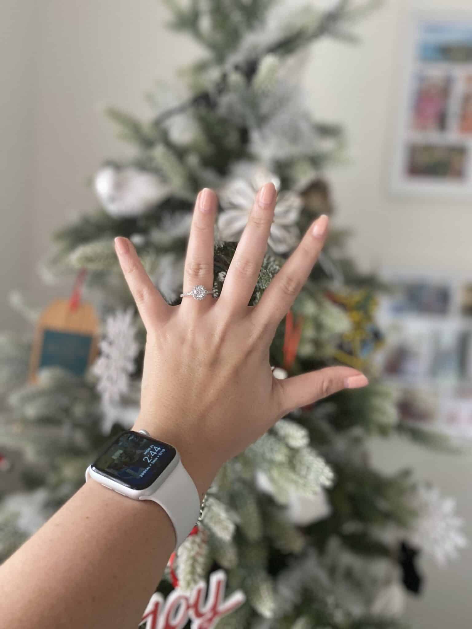 person holds out hand in front of christmas tree wearing apple watch with new engagement ring on hand